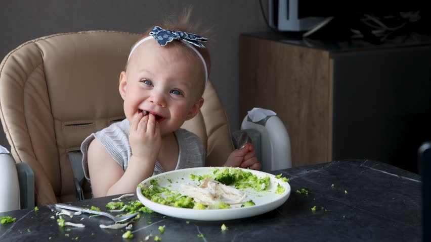 Funny messy baby girl eating food with spoon at table, adorable toddler learning to feed herself, cute child mealtime chaos, candid moment of childhood