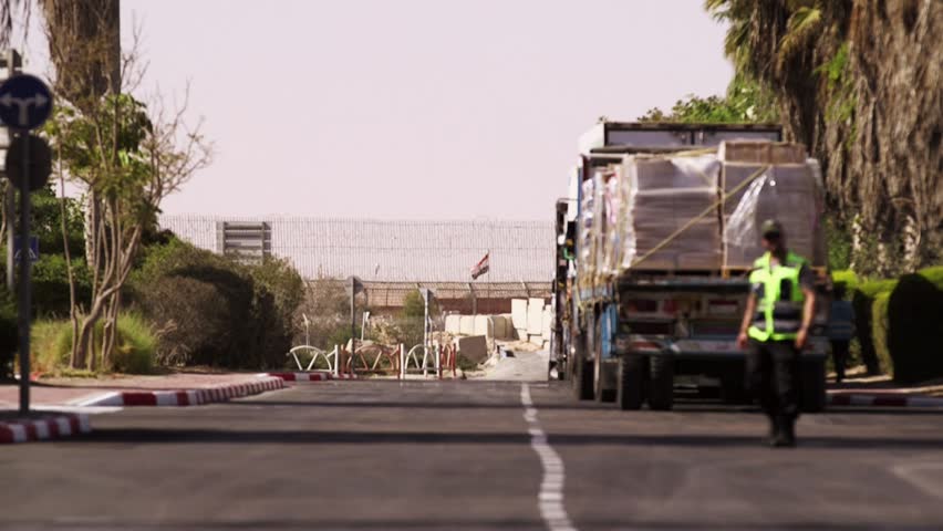 An extreme telephoto clip of the Israeli Gaza border fence and an Egyptian flag, with an aid truck and some workers in the unfocused foreground. none are recognizable.