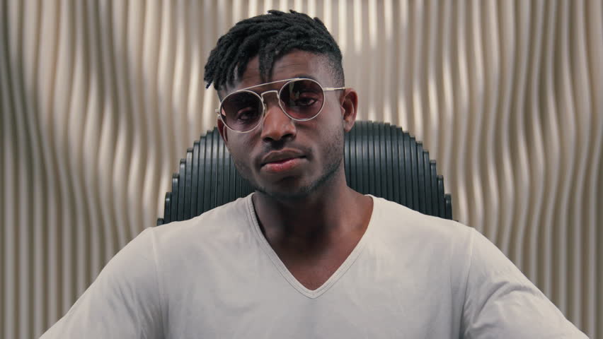 Portrait African American guy man ethnic male putting on sunglasses glasses wearing looking at camera sitting in futuristic white studio futurism innovation stylish fashion posing indoors sit in chair