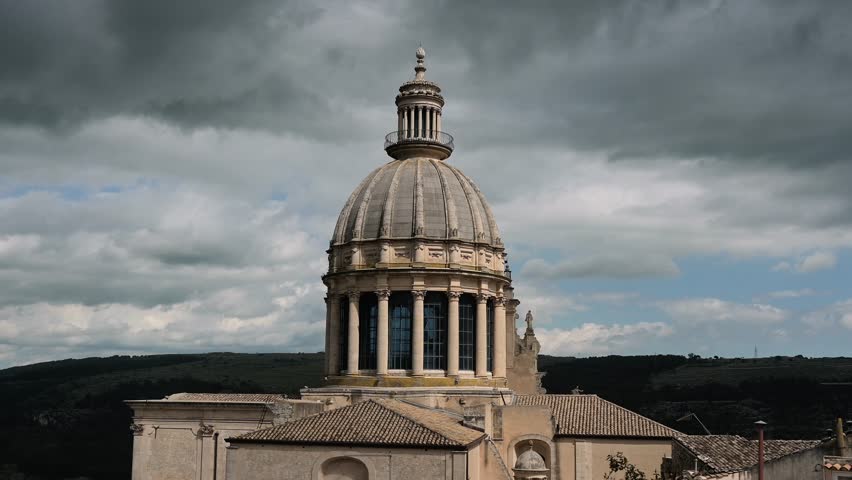 Prominent dome of San Giorgio cathedral in Ragusa Ibla stands tall above tiled rooftops, set against a dramatic sky with heavy, layered clouds.