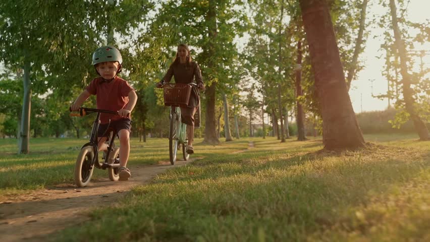 Young playful boy wearing helmet enjoys a fun bike ride on a sunny day, followed by his smiling mother through a green park trail. Mom and son riding bikes together in nature. Slow motion