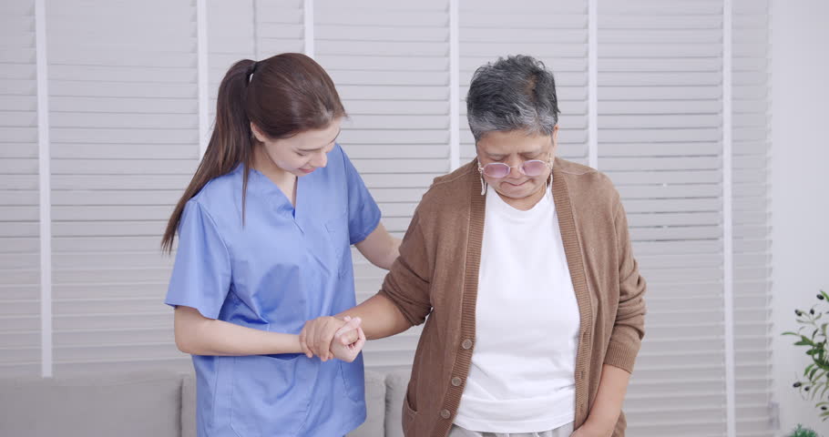 Slowmotion moment of Caucasian female physiotherapist gently supporting Asian senior woman walking step by step for balance and leg strength rehabilitation training during elderly care session