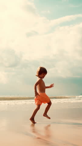 Vertical shot of Happy smiling kid 5 years old running along beach in water slow motion. Boy with long hair in pink swimming trunks runs cheerfully along beach in setting sun. Concept beach holiday
