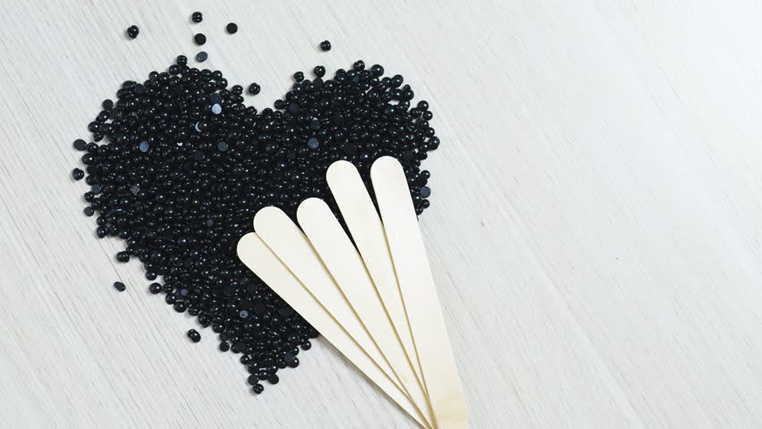 Black hard wax beans forming heart shape on rustic wooden surface, wooden spatulas nearby, representing beauty, hair removal, and skincare preparation