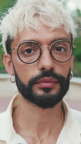 Vertical video, Portrait of a stylish young man with blond hair, beard and glasses in an urban setting. He looks directly at the camera with a serious expression.