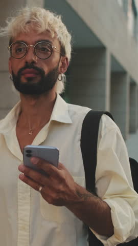 Vertical video, A young man with bleached blond hair and a beard smiles while walking down a city sidewalk and looking at his phone.