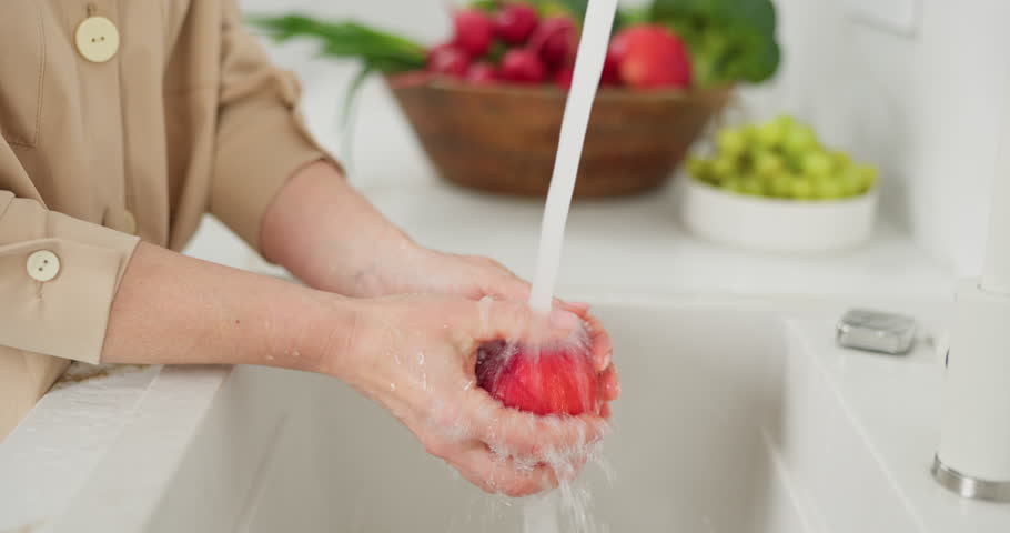 Slow-motion footage of womans hands wash red peach under the pressure of flowing water in the white kitchen, a basket with radishes, broccoli and onions in the background, washed green grapes in a cup