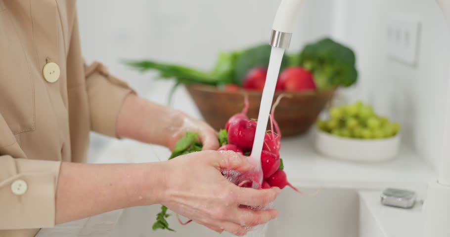 Slow-motion footage of woman's hands are washed a bunch of red radishes under the pressure of flowing water in the white kitchen, a basket with tomato, broccoli and onions in the background, grapes