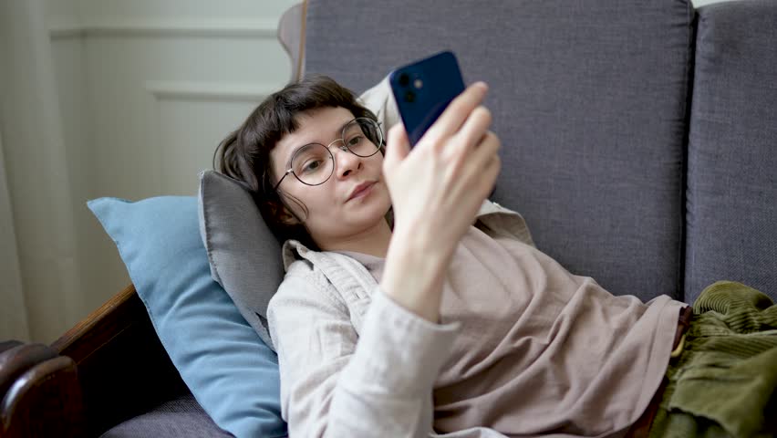 Relaxed woman browsing social media on smartphone while lounging on sofa, absorbed in digital web feed. Device dependency, endless scrolling habit, internet downtime, modern leisure, lonely isolation