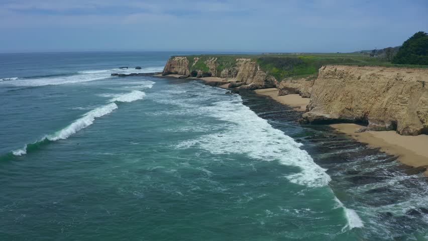 Aerial view of coastal cliffs and sandy beach in Davenport, California. Ocean waves roll onto shore near rugged rock formations and cypress trees. Perfect for travel, nature, and scenic coastline
