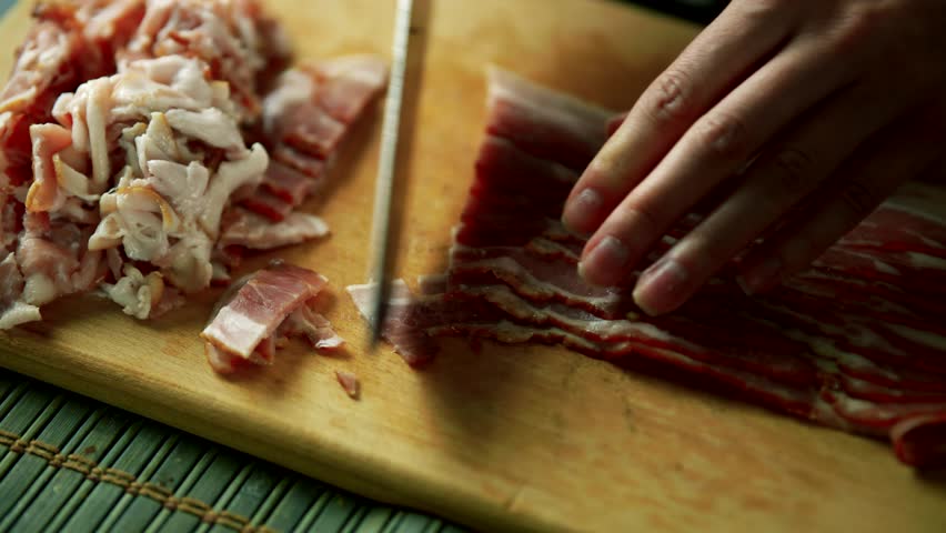 person slicing raw bacon on a wooden cutting board. Meat and a knife food preparation. Chef slicing bacon for pasta carbonara. Slice strips of raw bacon on a wooden cutting board. Savory dish bacon