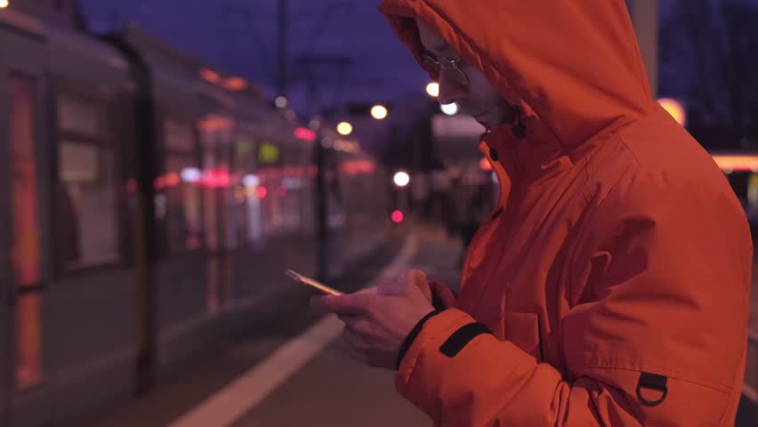 young man in orange jacket scrolling smartphone, browsing phone, surprised by online news, surfing on phone in city, wasting time, casual clothing, emotional face, screen addiction, daily routine
