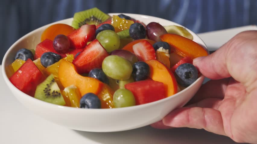 a full white bowl with fresh fruit piece salad is placed by a human hand on the table for a healthy meal, zoom