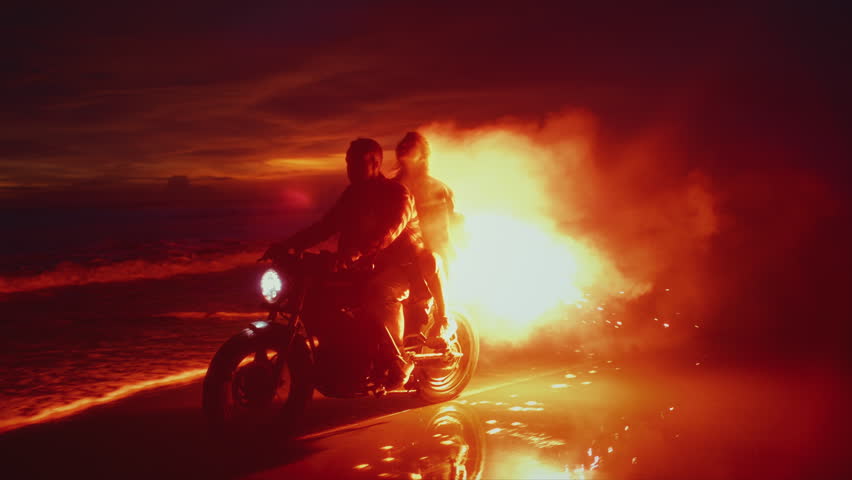 Two young couples riding on vintage motorcycles with red burning signal flares after sunset on beach, slow motion