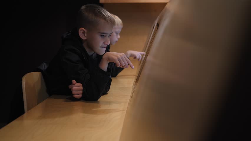 Two children are sitting in the library, in front of a electronic tablet with a touch screen. They are reading a book or article from the tablet, which is mounted on the wall of the modern library. - Powered by Shutterstock - Get 15% off with code: PIKWIZARD15