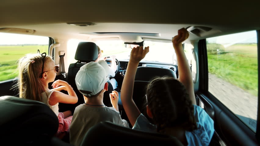 Mother takes her children by car to nature on a sunny summer day along a country road. The children are having fun in the back seat, singing and raising their hands, dancing to the music