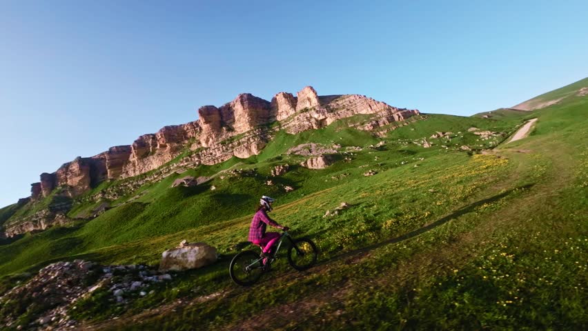 Young athlete cyclist in helmet and mask doing wheelie trick riding on rear wheel and pedaling in mountainous area at sunset. Aerial view shooting on fpv drone.