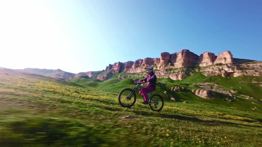 Young athlete cyclist in helmet and mask doing wheelie trick riding on rear wheel and pedaling in mountainous area at sunset. Aerial view shooting on fpv drone.