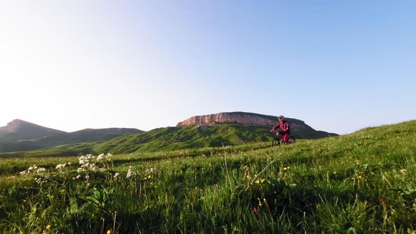 Young athlete cyclist in helmet and mask doing wheelie trick riding on rear wheel and pedaling in mountainous area at sunset. Aerial view shooting on fpv drone.