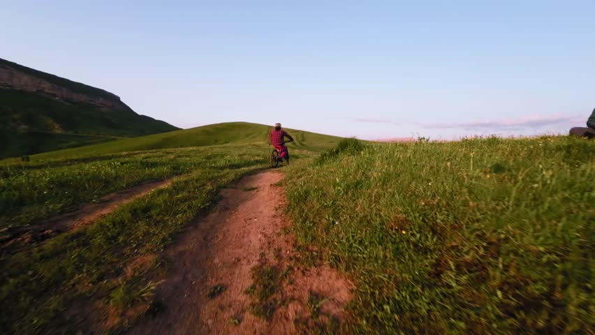 Young athlete cyclist in helmet and mask doing wheelie trick riding on rear wheel and pedaling in mountainous area at sunset. Aerial view shooting on fpv drone.