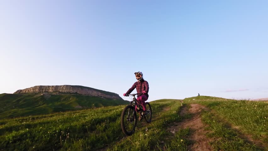 Young athlete cyclist in helmet and mask doing wheelie trick riding on rear wheel and pedaling in mountainous area at sunset. Aerial view shooting on fpv drone.