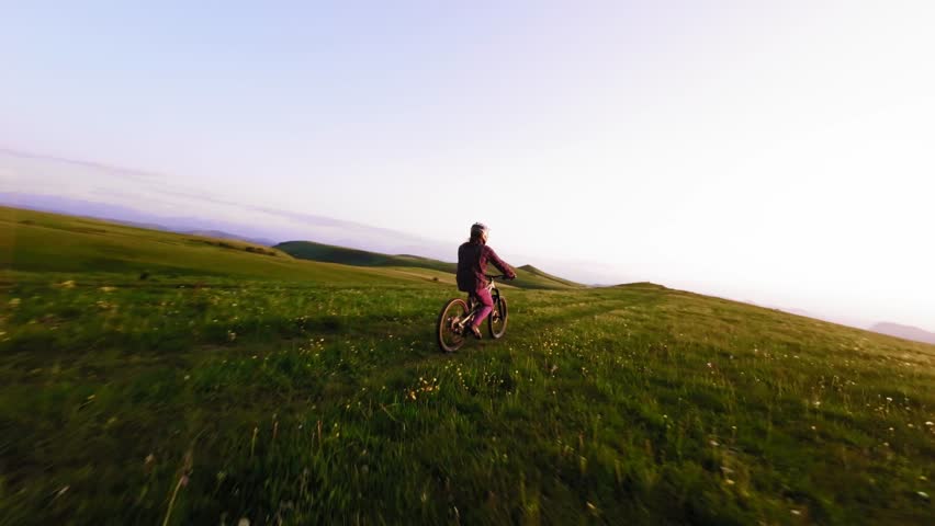 Young athlete cyclist in helmet and mask doing wheelie trick riding on rear wheel and pedaling in mountainous area at sunset. Aerial view shooting on fpv drone.