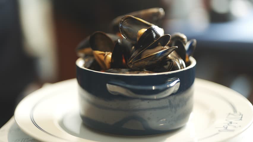 A blue bowl filled with mussels is placed on a white plate. The bowl is filled with a variety of mussels, including some that are larger and some that are smaller