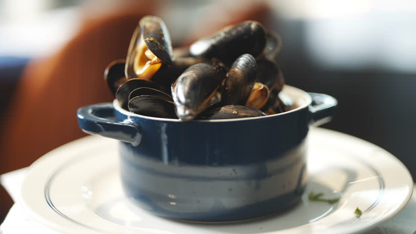 A blue ceramic bowl filled with mussels is placed on a white plate. The bowl is filled with a variety of mussels, including some that are larger and some that are smaller