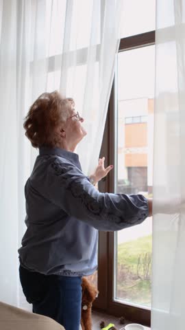 Senior woman standing by window at home, opening curtains to look outside and check weather, indoor, lifestyle