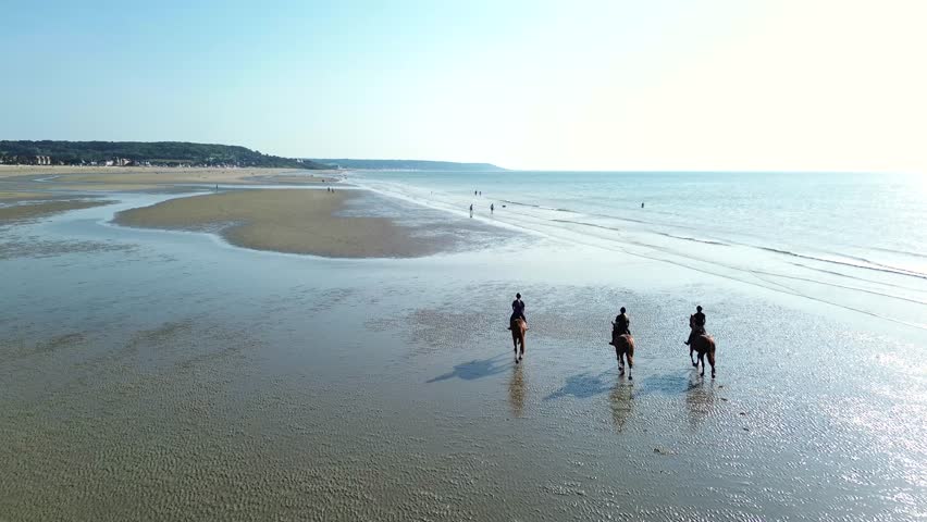 Drone shots of riders galloping at full speed across the reflective shoreline of Deauville beach. The sun glistens on the water as hooves kick up spray,