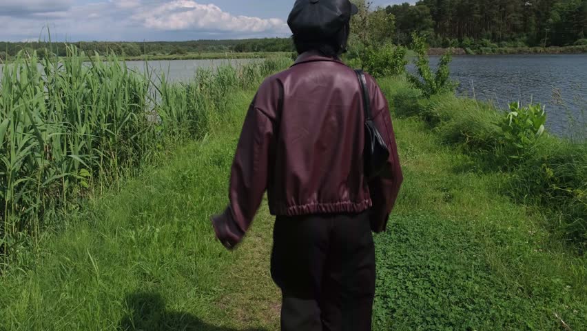 A woman in a leather jacket walks on a narrow path between two rivers or lakes, surrounded by lush greenery and summer nature.