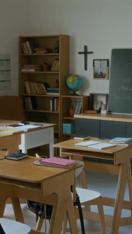 Vertical no people shot of empty classroom with single school desks and chairs in modern religious school with Cristian cross on wall