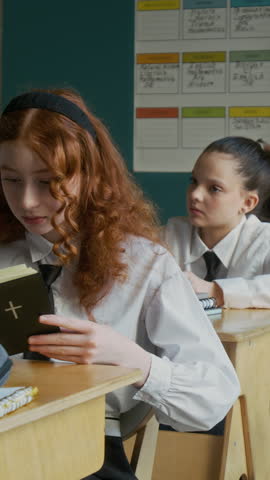 Vertical medium shot of focused teen Caucasian girl in white uniform reading Bible sitting at desk during class in Christian school