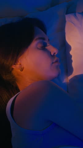 Vertical shot of brunette young woman in comfy pajamas lying in bed and sleeping peacefully while bedroom illumed by deep blue and golden lights