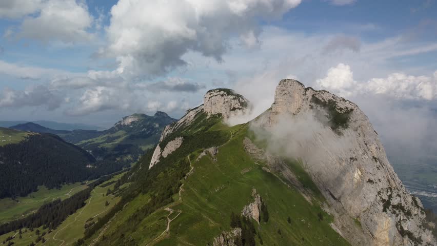 Aerial video of mountain ridge with light clouds drifting across rocky peaks and green slopes, revealing hiking trail and panoramic alpine landscape under dynamic sky