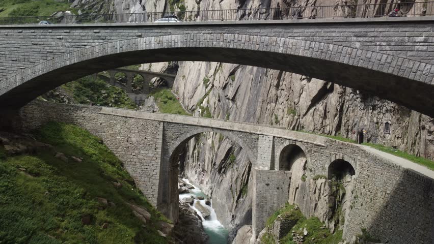 Aerial video of historic arched stone bridges crossing a deep mountain gorge with turquoise river, surrounded by steep cliffs and rugged alpine terrain.