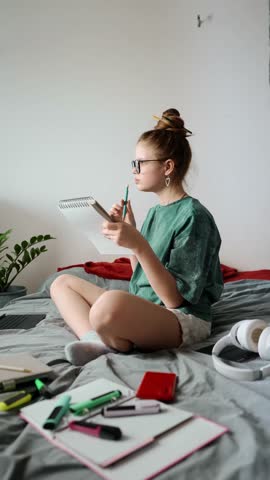 Student girl with glasses and cozy outfit studying at home, writing in notebook, surrounded by stationery, enjoying remote education in relaxed bedroom setup.