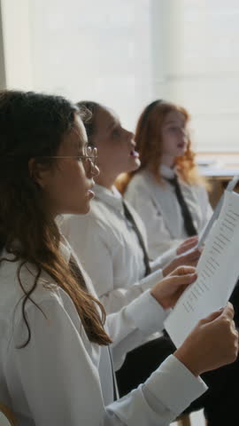 Vertical side view shot of motivated multiethnic student girls in white uniforms standing in circle in classroom and singing in choir holding lyrics in hands