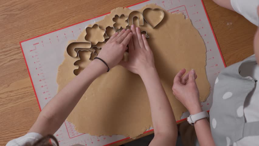 Hands carefully press various metal cookie cutters into fresh gingerbread dough. Festive holiday shapes, including a teddy bear, star, and heart, are being crafted on a silicone baking mat.
