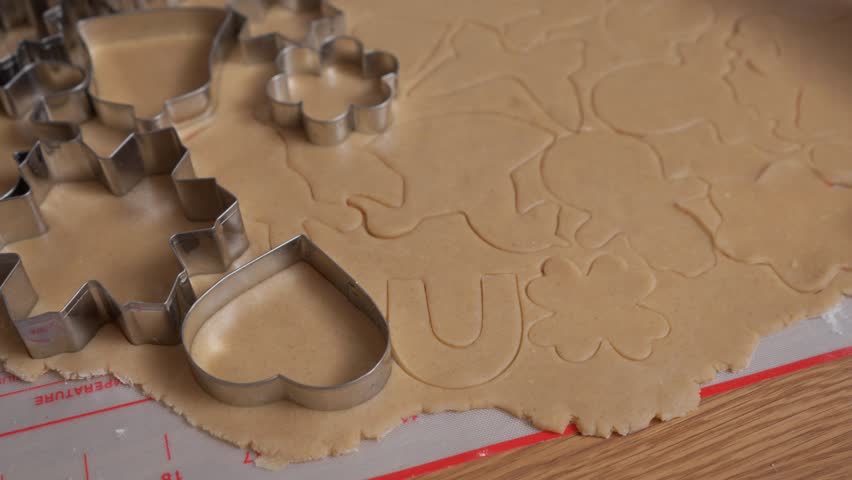 An intricate pattern of holiday cookie shapes covers a sheet of raw gingerbread dough. The detailed texture of the pastry is visible where hands have pressed in various metal cutters.
