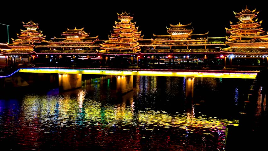 Experience the breathtaking transformation of the wind and rain bridge at night in Qiandongnan Guizhou Province, showcasing stunning reflections in the water. Watch as lights dance across the scenery.
