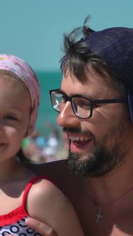 Father and little daughter look at camera and laugh on background of beach with blue sea. Summer vacation travel holiday. People outdoors on seascape. Vertical shot