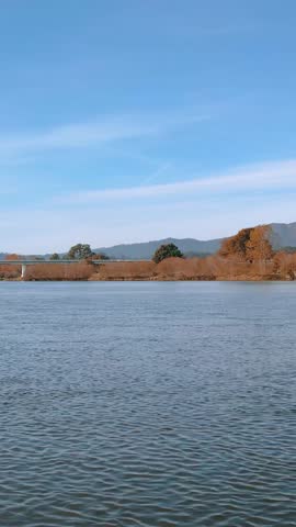Serene riverscape with the bridge visible across the water and distant wooded hills under a pale blue autumn sky.
