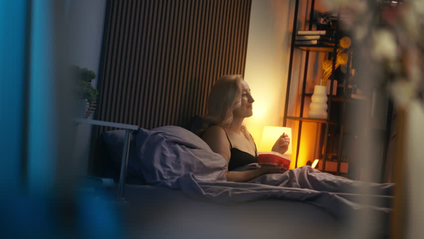 Happy middle-aged woman eating ice cream and watching movie in bed, evening rest
