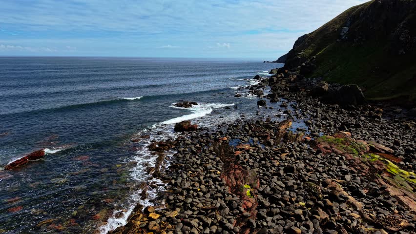 Waves rolling onto rocky shore of North Sea in Scotland. Swells crashing against boulders along coastal landscape. Tides surging over stones near steep grassy cliffs