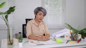 Senior Asian female architect sketching house plans at bright workspace with green plants and white blinds - Powered by Shutterstock - Get 15% off with code: PIKWIZARD15