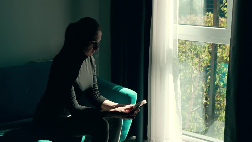 Woman holding UK cash and planning finances near bright window in modern hotel room. Counting British currency while sitting in teal chair with sunlight streaming through curtain. Managing budget with