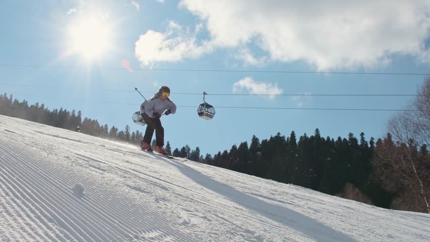 Young woman child skier with friends riding downhill on groomed snowy slope under bright winter sun with ski lift chairs and forest background in mountain scene