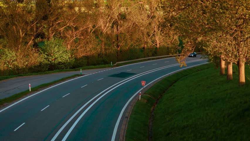 Cars driving on country highway curve in evening sunlight. Passenger vehicles moving through rural bend lined with spring trees. Autos traveling along scenic asphalt road bordered by fresh greenery