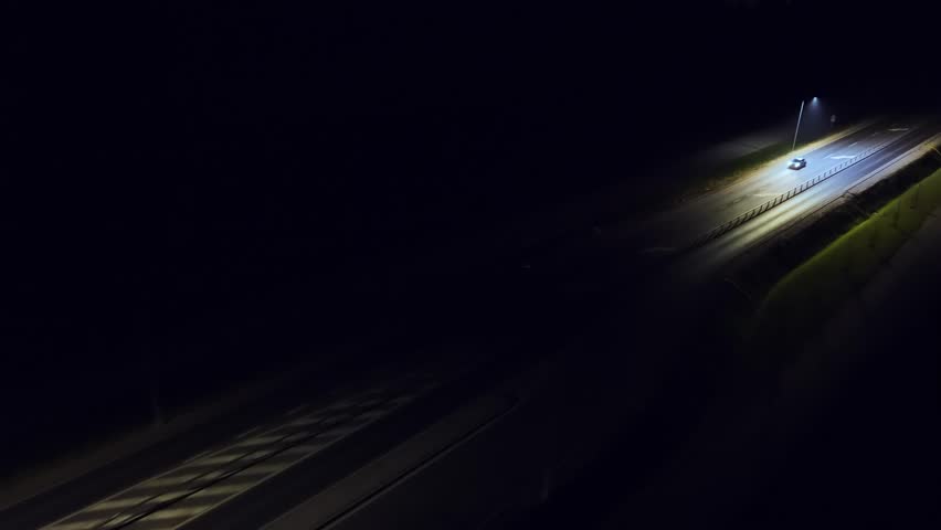 Car driving at night with headlights on along road lit by streetlights. Vehicle moving through darkness, beams illuminating way. Auto traveling along nighttime street, lights guiding direction. Sedan